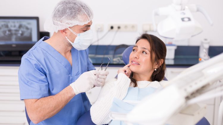 Woman in discomfort at a dental clinic, talking to a dentist holding tools.